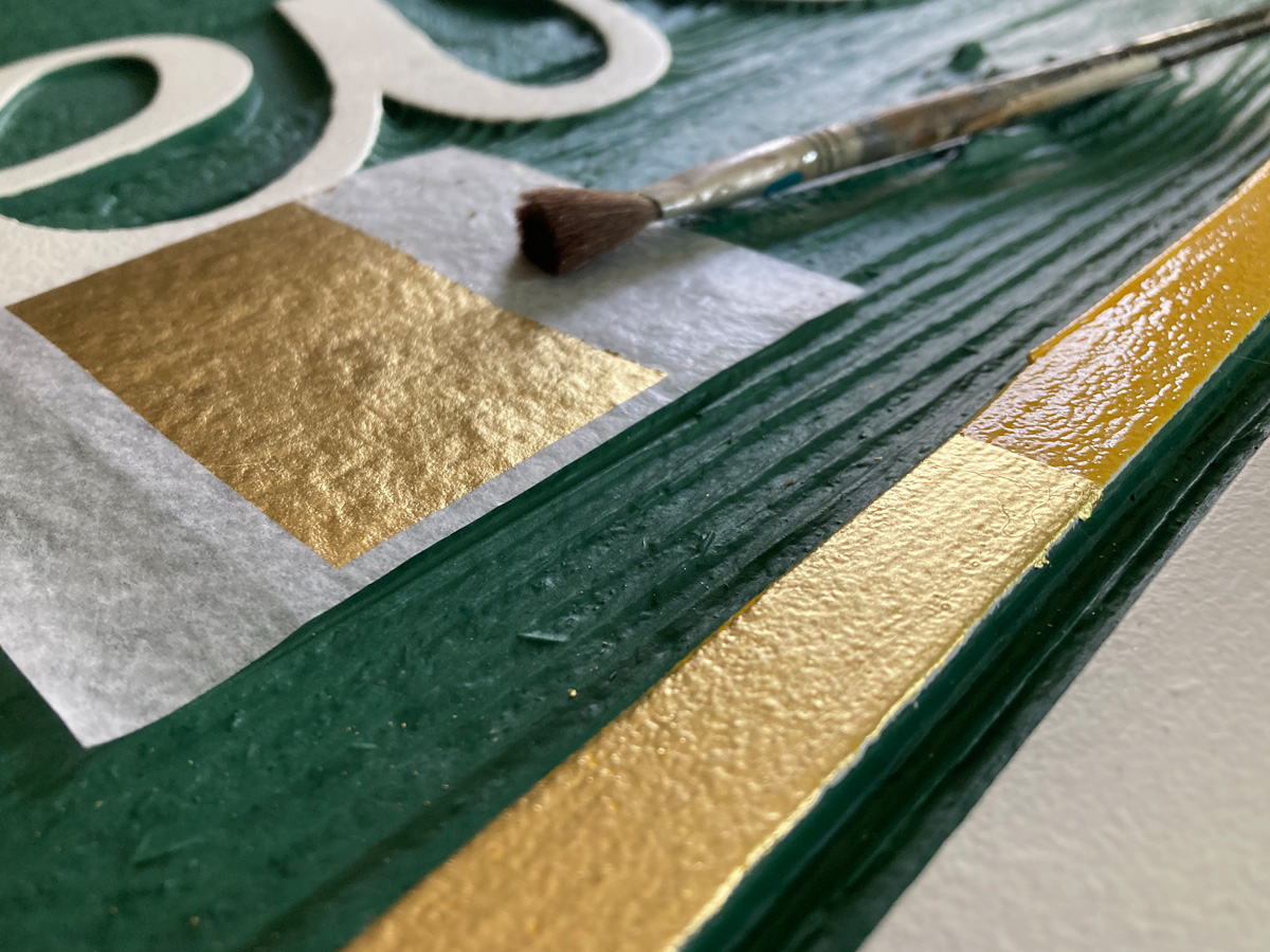 Traditional gold leaf being applied by hand to a carved green sign at Asheville Signworks